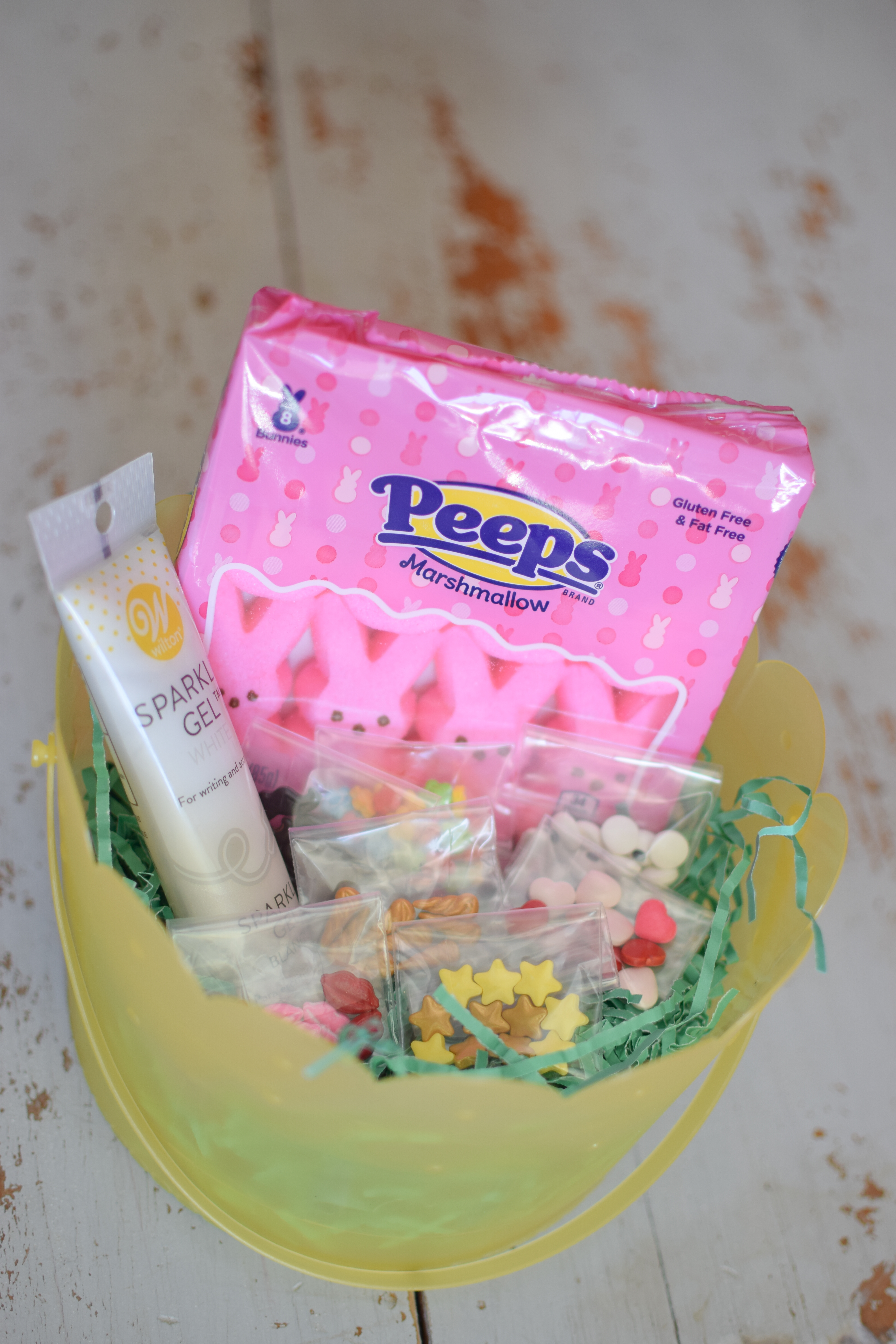 Peeps, assorted candy sprinkles and icing gel in a plastic yellow Easter bucket with shredded green paper grass